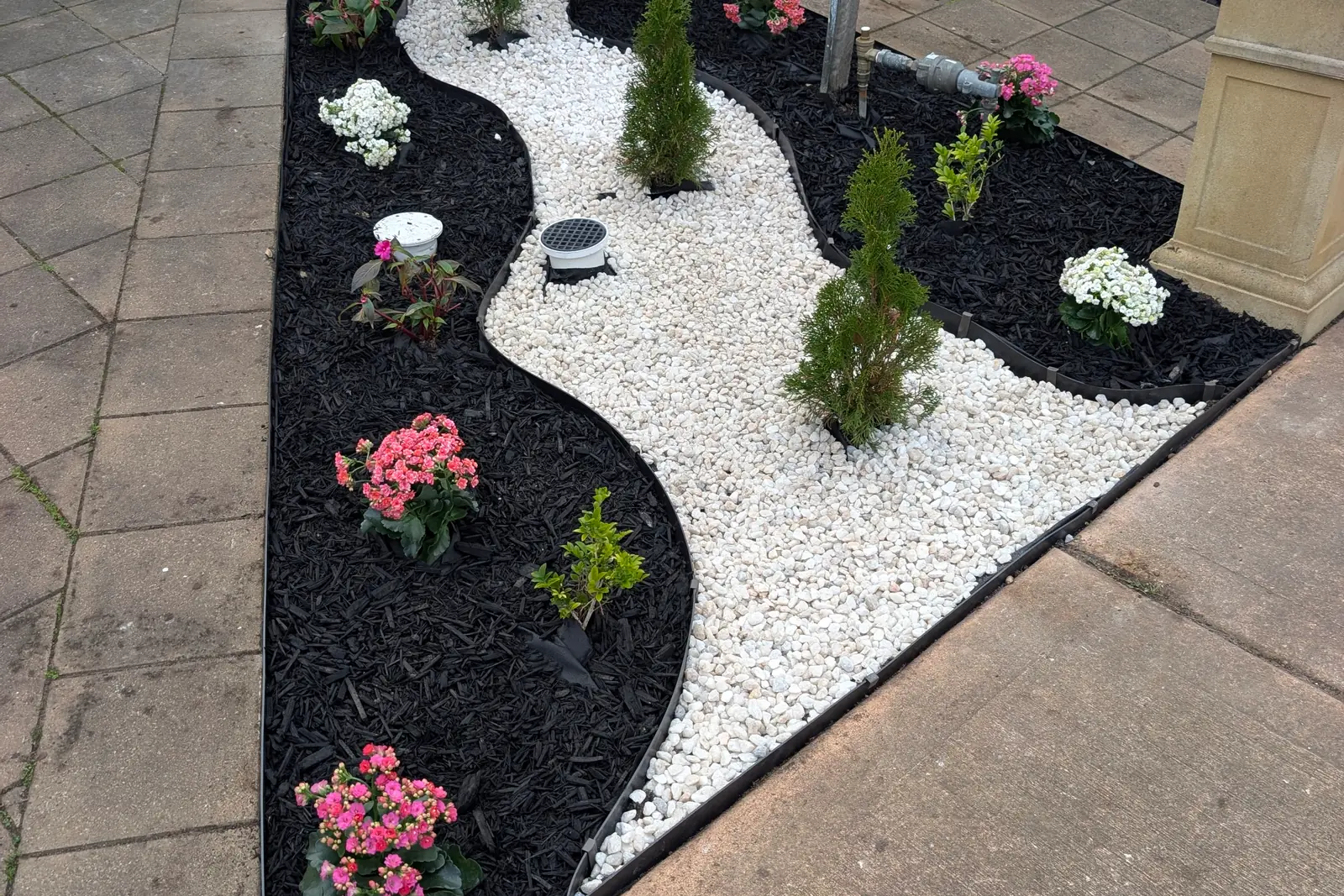Front garden bed with a curving band of white pebbles between black mulch, black edging and young shrubs and flowering plants