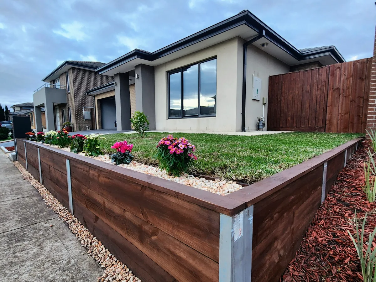 After: level lawn held by a dark-stained timber sleeper retaining wall with galvanised posts, white-pebble trim, flowers and red mulch